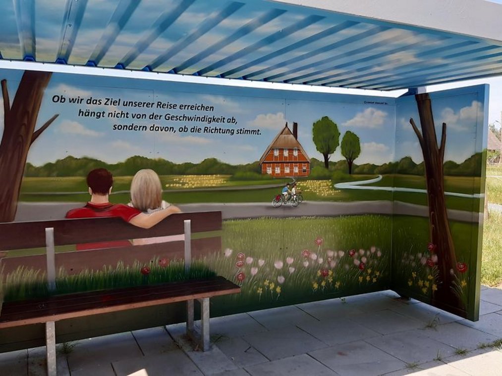 Buswartehäuschen mit gemaltem Landschaftsmotiv, Bank und Spruch über das Reiseziel