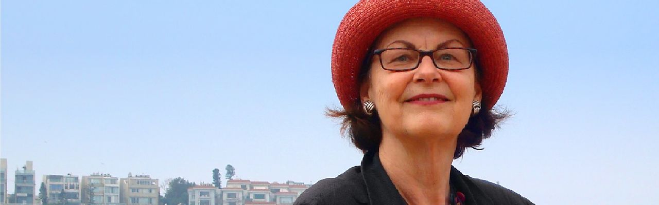 Lächelnde Frau mit rotem Hut und Brille vor Häusern und blauem Himmel