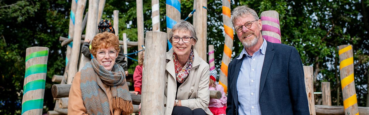 Zwei Frauen und ein Mann vor einem bunten Klettergerüst auf einem Spielplatz